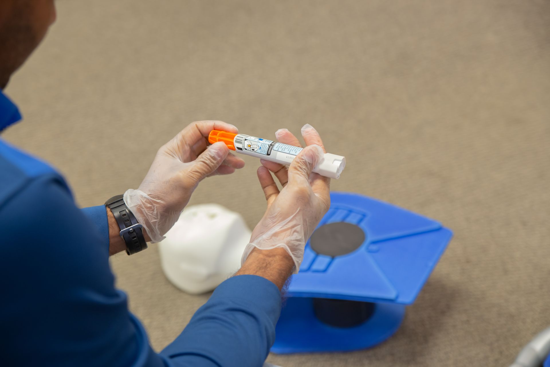 A First Aid and CPR level HCP trainee with gloves on holding an EpiPen with a blue mannequin / dummy for training behind it 