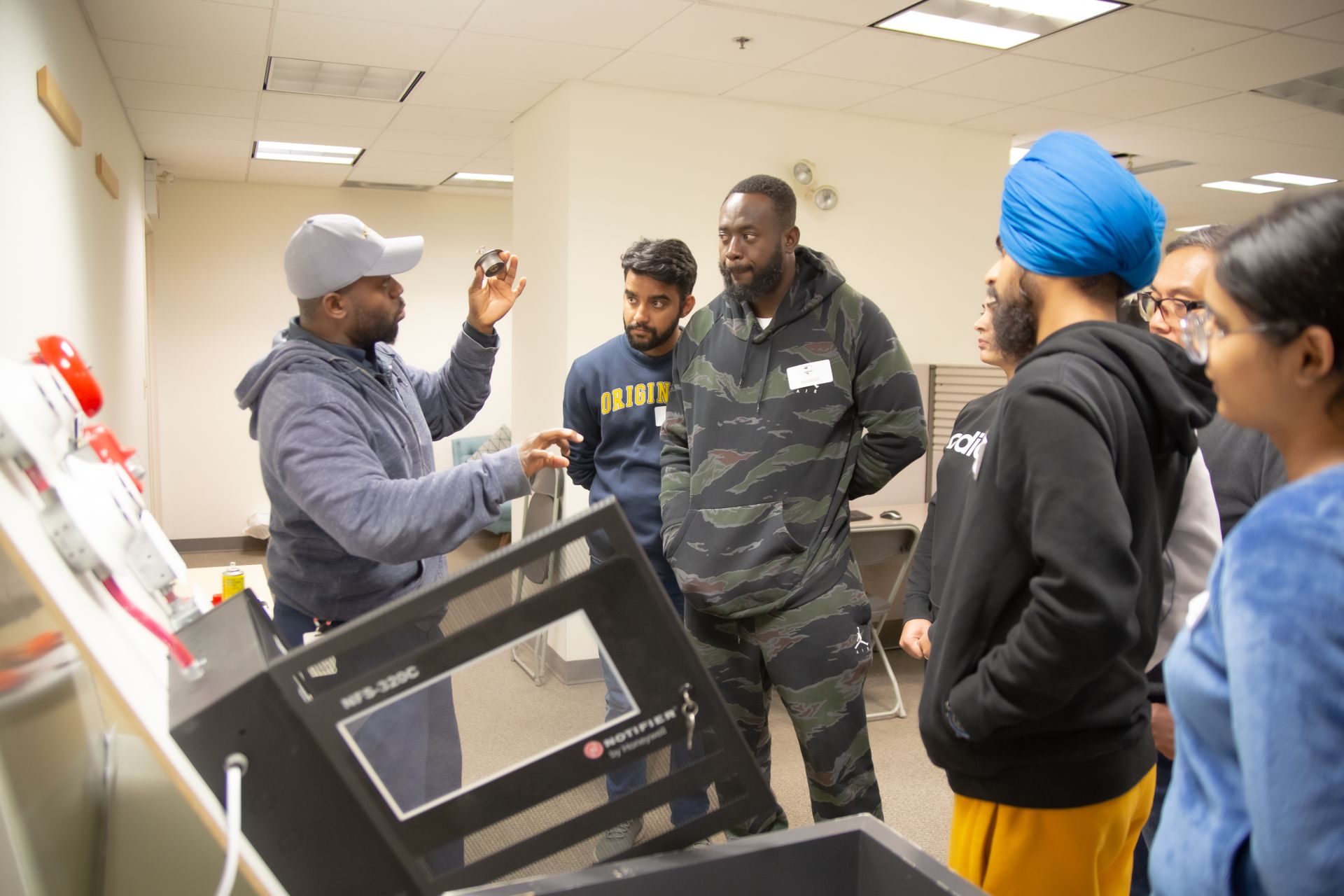 A class of Fire Alarm training students standing in front of a Flex Point Academy instructor who is standing next to a fire alarm panel for education purposes