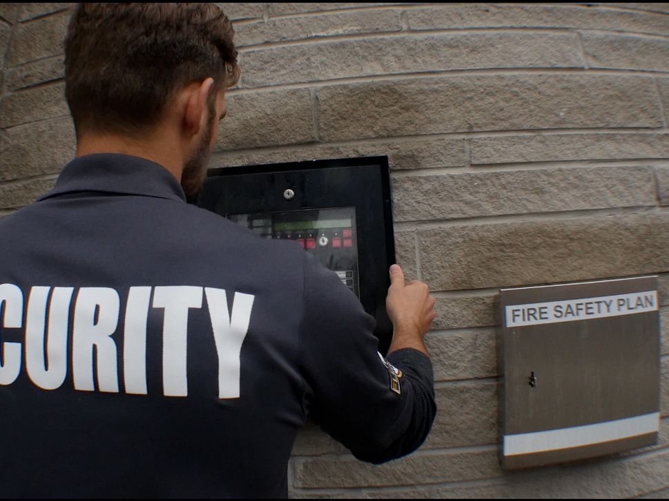 A male Flex Point Security guard about to open a Fire Alarm Panel mounted to a wall for review