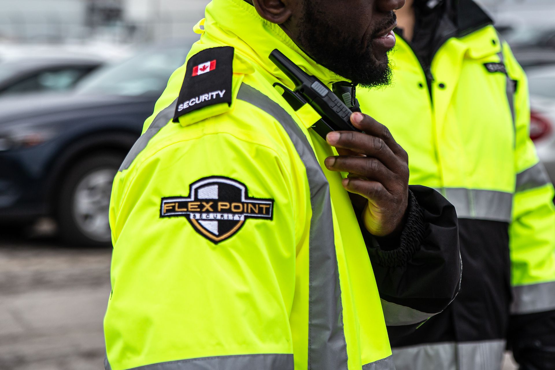 A male Flex Point Security guard wearing a branded yellow jacket reaching for a walkie talkie that is on his shoulder