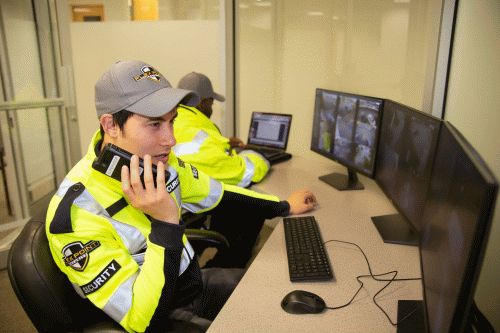 Two male Flex Point Security guards monitoring surveillance monitors while one of them speaks with someone using a walkie talkie