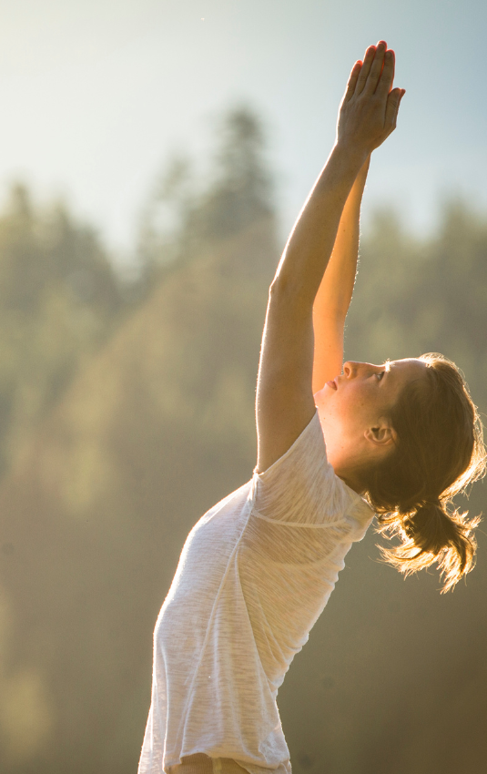A woman stands with hands raised overhead, fingers pressed together. Her gaze is turned toward her hands as soft sunlight radiates down. She is wearing a white shirt and has brown hair pulled back into a ponytail. The background is in soft focus with evergreen trees in the distance.