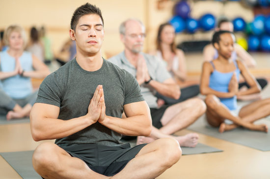 A group of people sit cross-legged on yoga mats in a studio, eyes closed in meditation, with a man in the foreground holding his hands together at his chest while others practice calmly behind him.