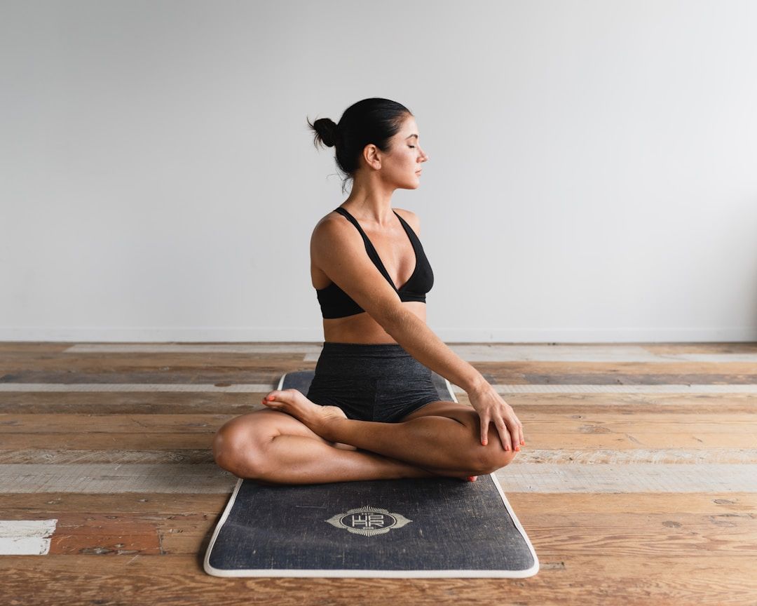 A yogi is shown sitting on their black and grey mat on a patched, wooden floor with old boards. The wall behind them is white. They are wearing a black sports bra and black, shorts pants and have their legs crossed in front of them, left foot resting on the right thigh. Their torso is twisted to the left, with the right hand gripping the left knee for stabilisation. 