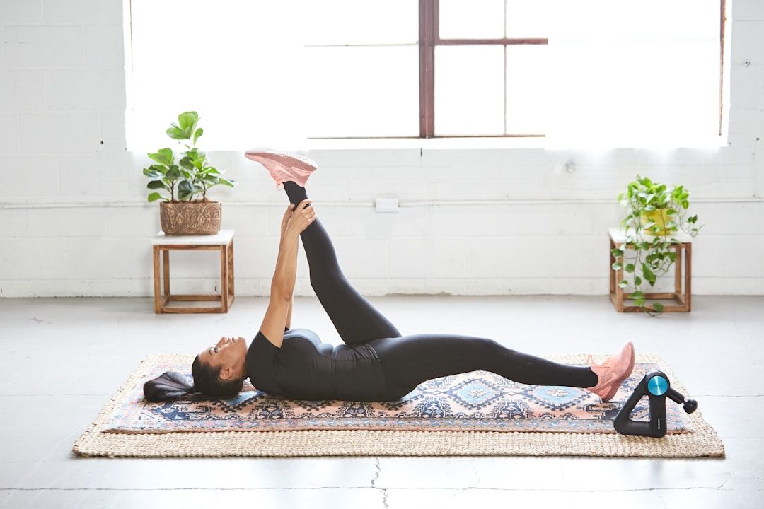 A woman sits on the ground in front of her laptop with her legs crossed. She's resting her hands on her knees facing upward, thumb and index fingers touching in a meditative pose. The woman is dressed in fitness clothes in earth tones, the background holds a large window front including an indoor palm tree and a yellow sofa.