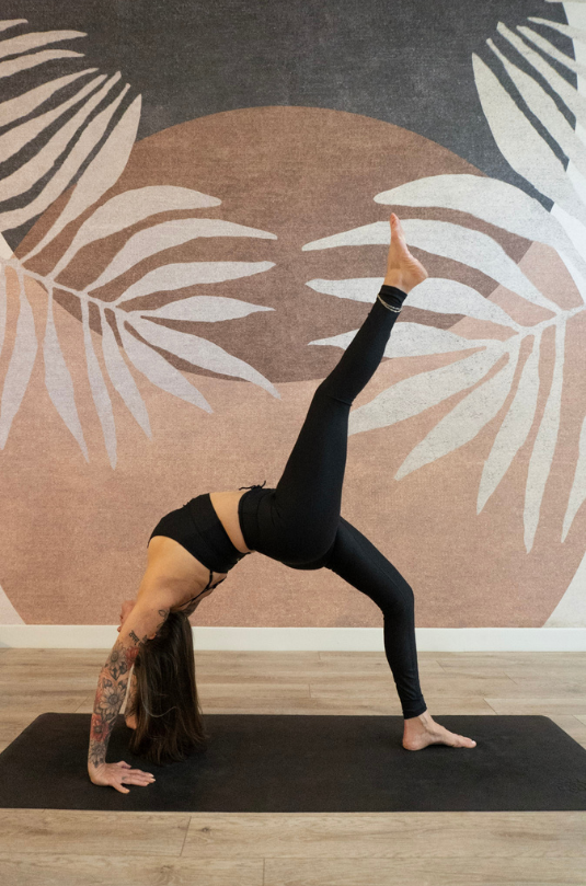 A yogi in black gym clothes sits on a black yoga mat in a studio with wooden floors. Their hands rest on their knees and their legs are crossed in front of them.