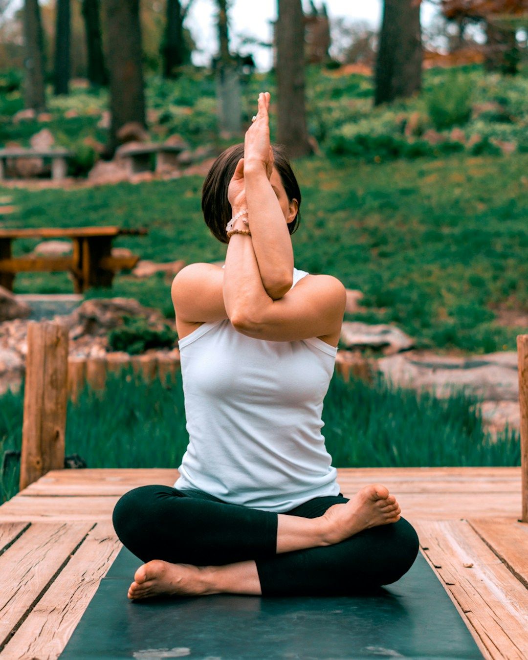 a woman sits in Lotus Pose with her right foot resting on top of the left knoww. The arms are raised in Eagle Pose, with the left arm wrapping underneath the right, palms touching each other in front of the face. The background is a serene nature spot with trees and foliage.