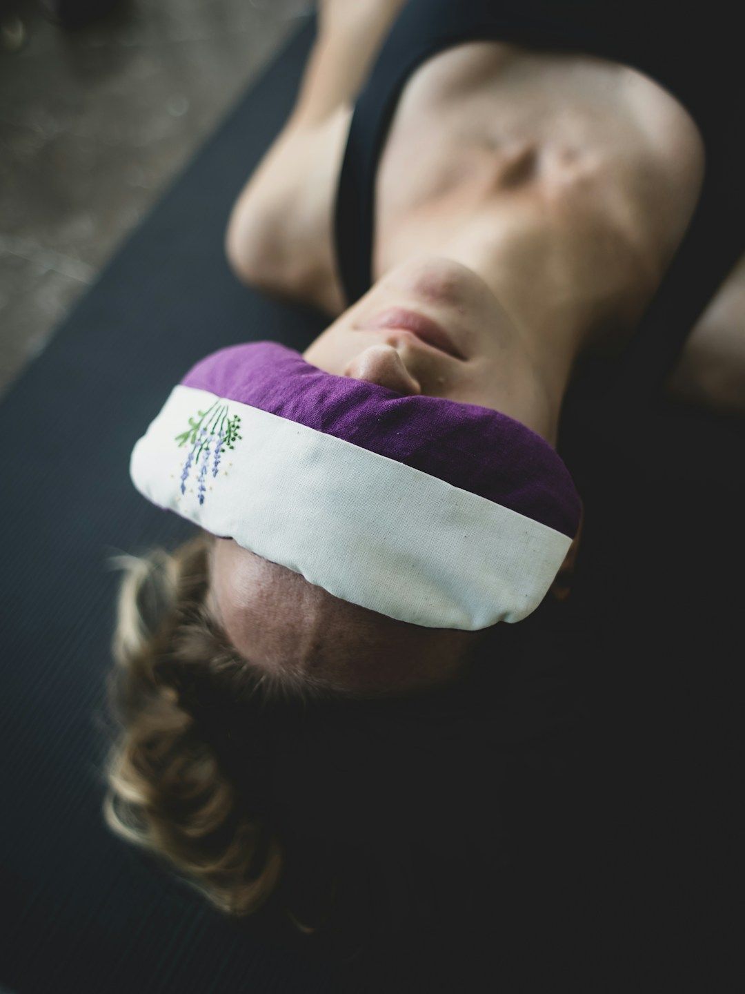 A woman is resting on a black yoga mat against black ground. She's got a lavender eyepillow over her face for ultimate rest.