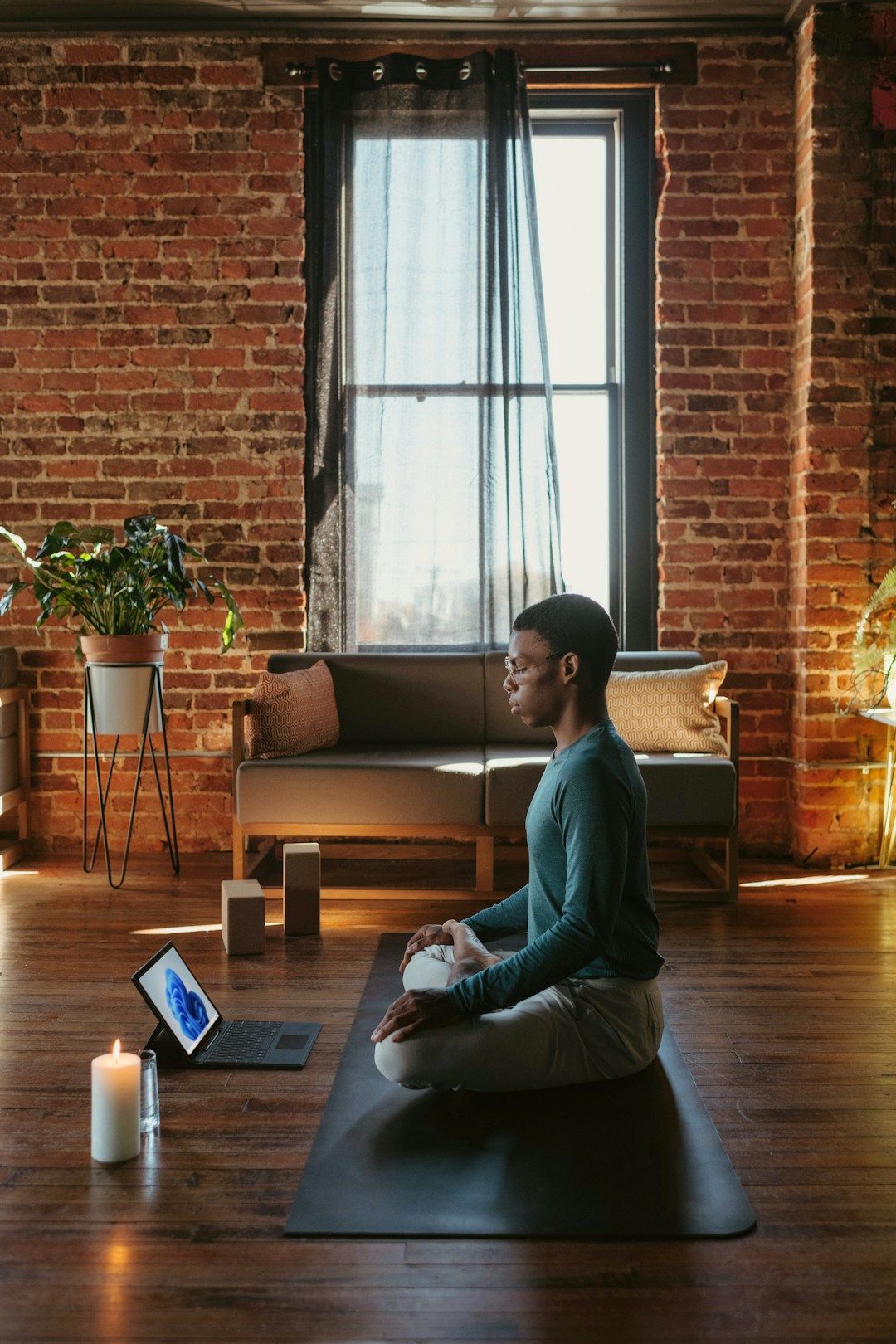 A woman sits on the ground in front of her laptop with her legs crossed. She has her left hand on the opposite knee in a stretch. She has curly hair and sits in front of a green plant. There are candles lit around her.