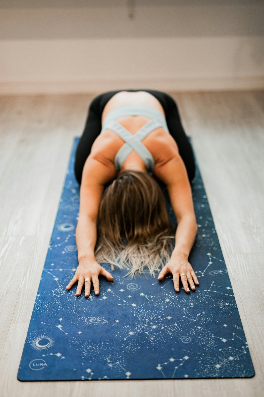 A yogi in a grey top adn black pants is shown resting in Child's Pose on their blue mat with a white solar system print. Her hands are strechted forward, while the yogi is sitting back on their heels.