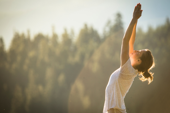 A woman stands in soft, golden morning light with a softened background of trees. She has her arms extended overhead and lifted into prayer pose, with her gaze upturned.