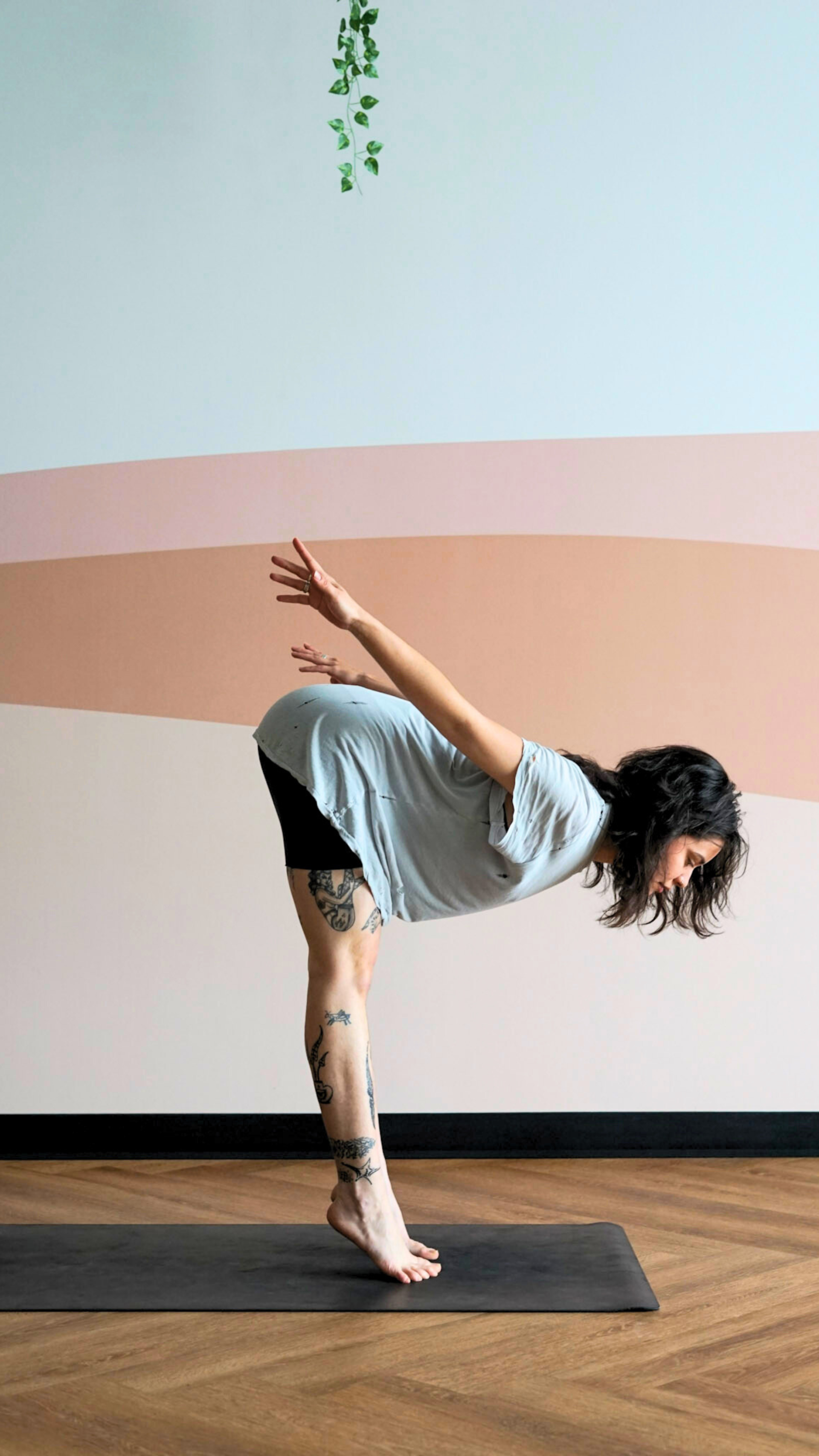 A yogi is shown in Diver's pose, balancing on thetips of their toes with the heels slightly raised, legs straight. Torso is bent forward at slightly more than 90 degrees and their arms are lifted behind their back. Their on a black yoga mat on wooden floor, with a waal with greenery and sand, old-rose stripes.