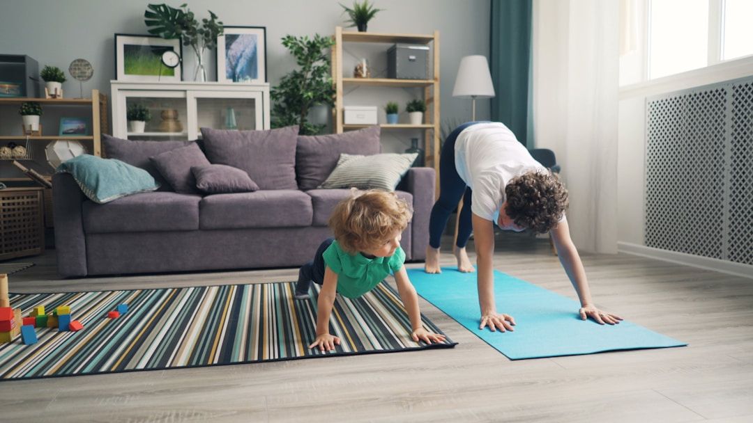 Two children are shown practicing yoga in a living room, one on a carpet, the other on a light blue yoga mat. They are in Plank Pose and Downward Dog. 