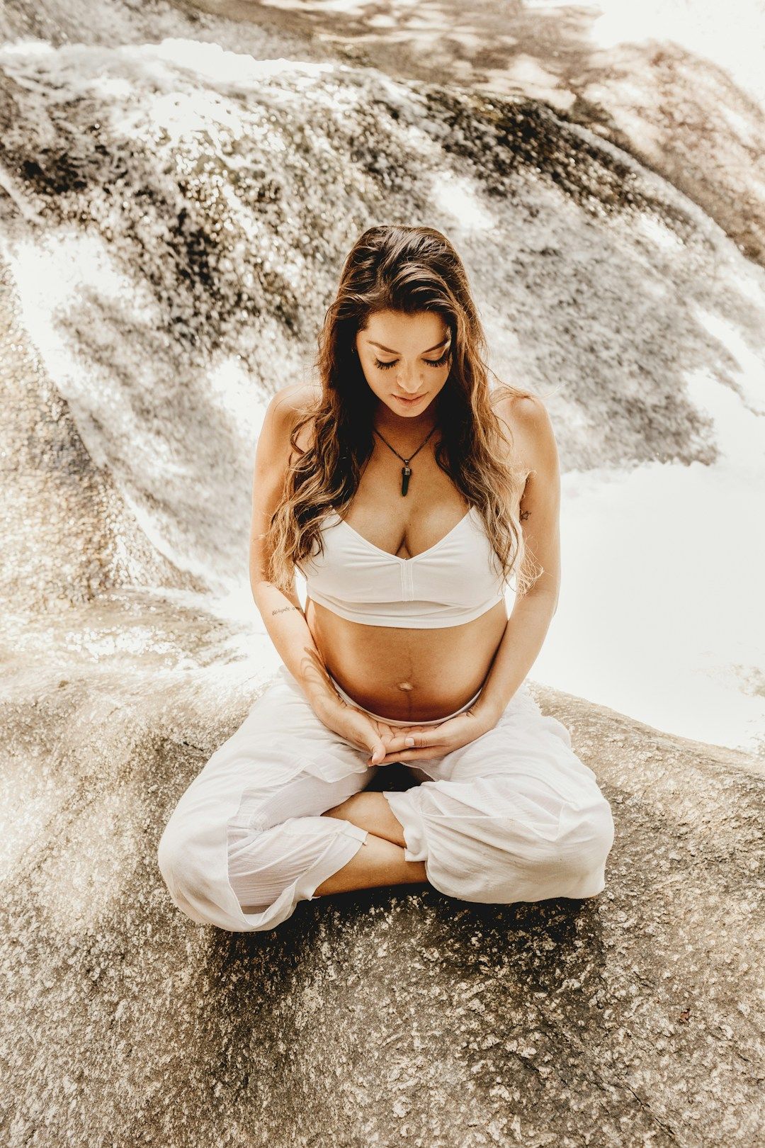 A pregnant woman with long, blonde waves falling open onto both shoulders is sitting on a rock in a water feature, legs crossed in front of her. She’s wearing white pants and a white sports bra, holding her belly with both hands. 