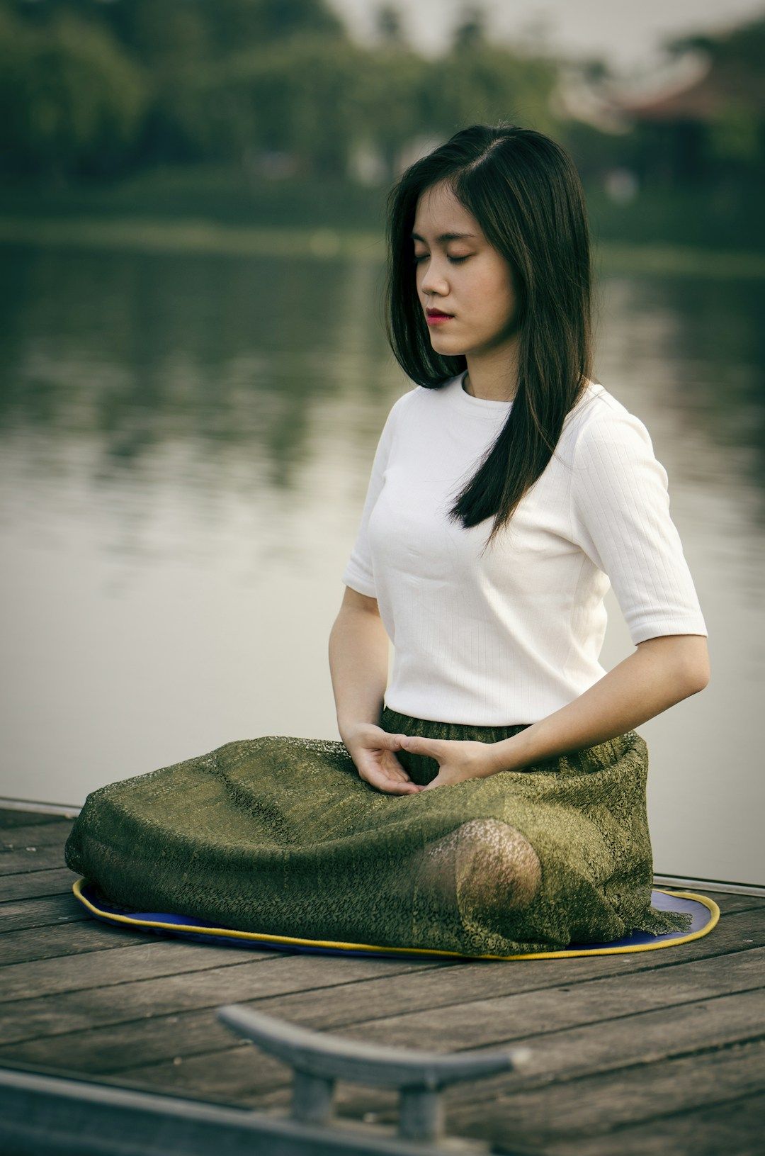 A woman with long, black hair that falls open onto her back and left shoulder, sits still in meditation on a wooden path above a still body of water. She’s wearing a green, long skirt, fully covering her legs and feet, and a white, half-arm sleeve shirt. Her hands are in a Mudra in her lap.