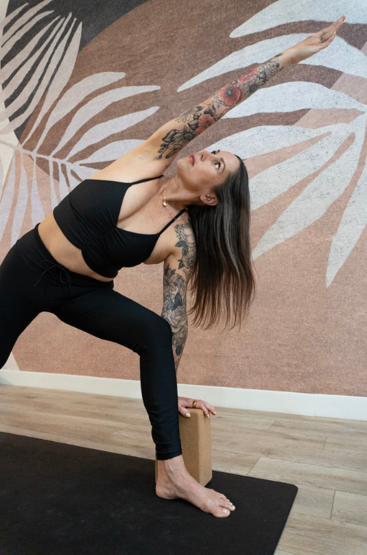 A person with long dark hair and tattoos on both arms is practicing yoga in a studio. They are performing a side stretch pose, reaching their right arm upward while supporting their left hand on a yoga block. The background features a neutral-colored wall with abstract leaf patterns. The person is wearing a black sports bra and leggings.