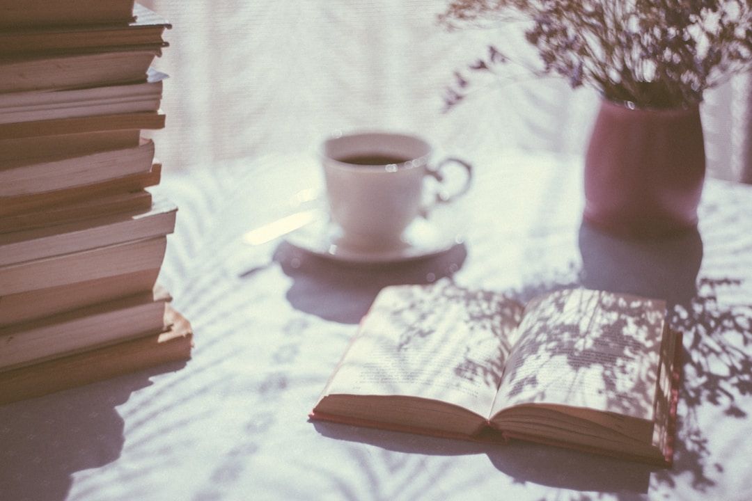 A book is shown on a table with a white table cloth, lying open. There's a bunch of books on the left, a white teacup, and a flower pot on the table. 