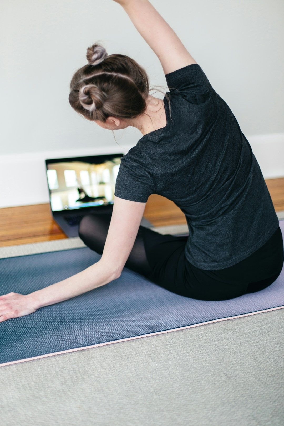A woman is seated on the floor her legs in front of her, her back facing the camera. The woman has her right arm lifted above her head and is bending her torso to the left. In front of her is a computer, demonstrating an online yoga class the woman is following.
