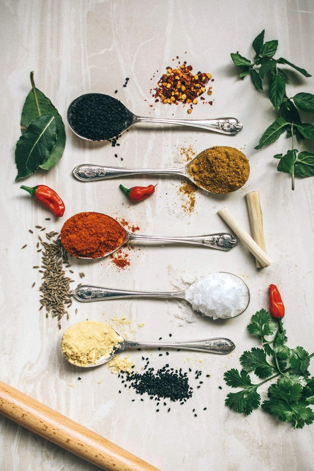 * A light wooden kitchen counter from above, showing several round silver spoons with different spice powders ranging in colours from black, to red, to yellow, to white. There are leaves, a few other spices, chilis, and a wooden stick.