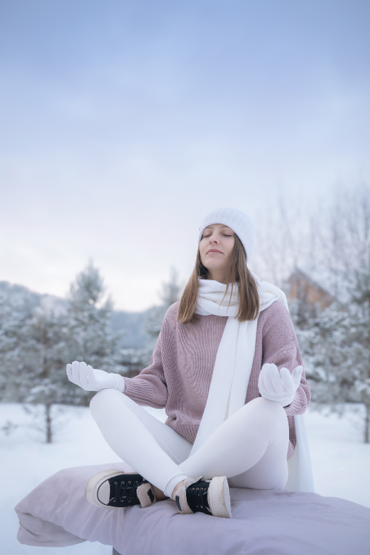 A woman sits cross-legged outdoors on a snowy day, meditating with her eyes closed. She is dressed warmly in a white hat, scarf, gloves, and leggings, along with a mauve sweater and black sneakers. Snow-covered trees and a soft winter sky create a calm, serene background.