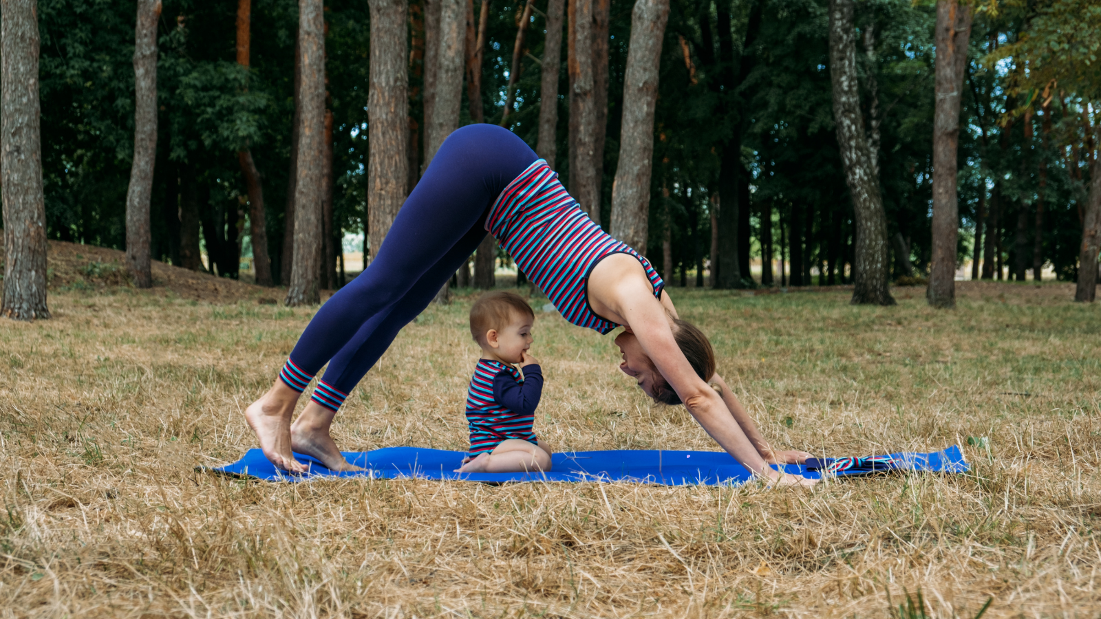 A woman does downward facing dog on her yoga mat in an outdoor setting while her child is underneath her. She is wearing a striped athletic top that matches her child's outfit.
