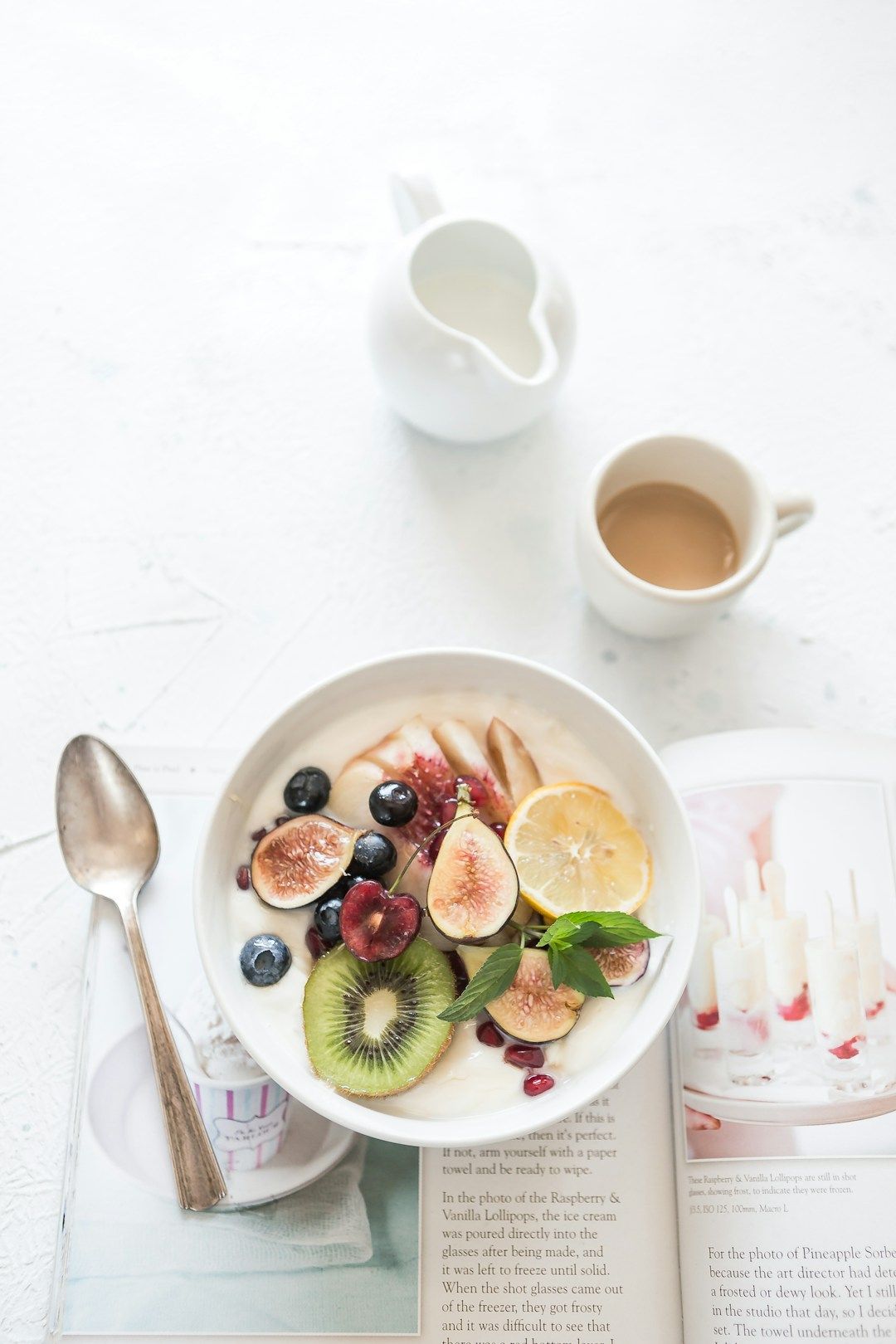 A kitchen counter is shown from above, a magazine is lying open on some recipes with a bowl with yoghurt and fresh fruits cut in standing on top. There’s also a white coffee cup and milk mug, and a silver spoon.