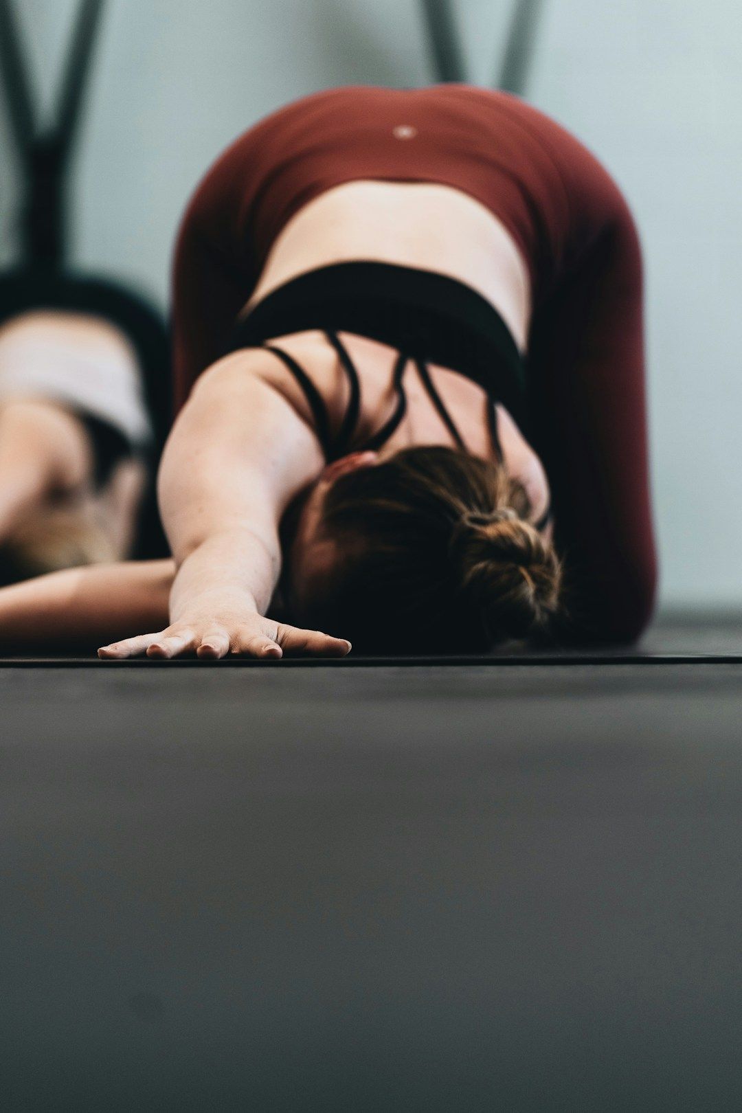 A woman with blonde hair in a bun is shown in Three the Needle Pose, kneeling with both hips lifted straight above the knees. The torso is bent forward with the right hand stretching forward and resting on the mat. The left arm is threaded underneath the torso, reaching far to the right. She’s wearing a black sports top and maroon yoga pants. 