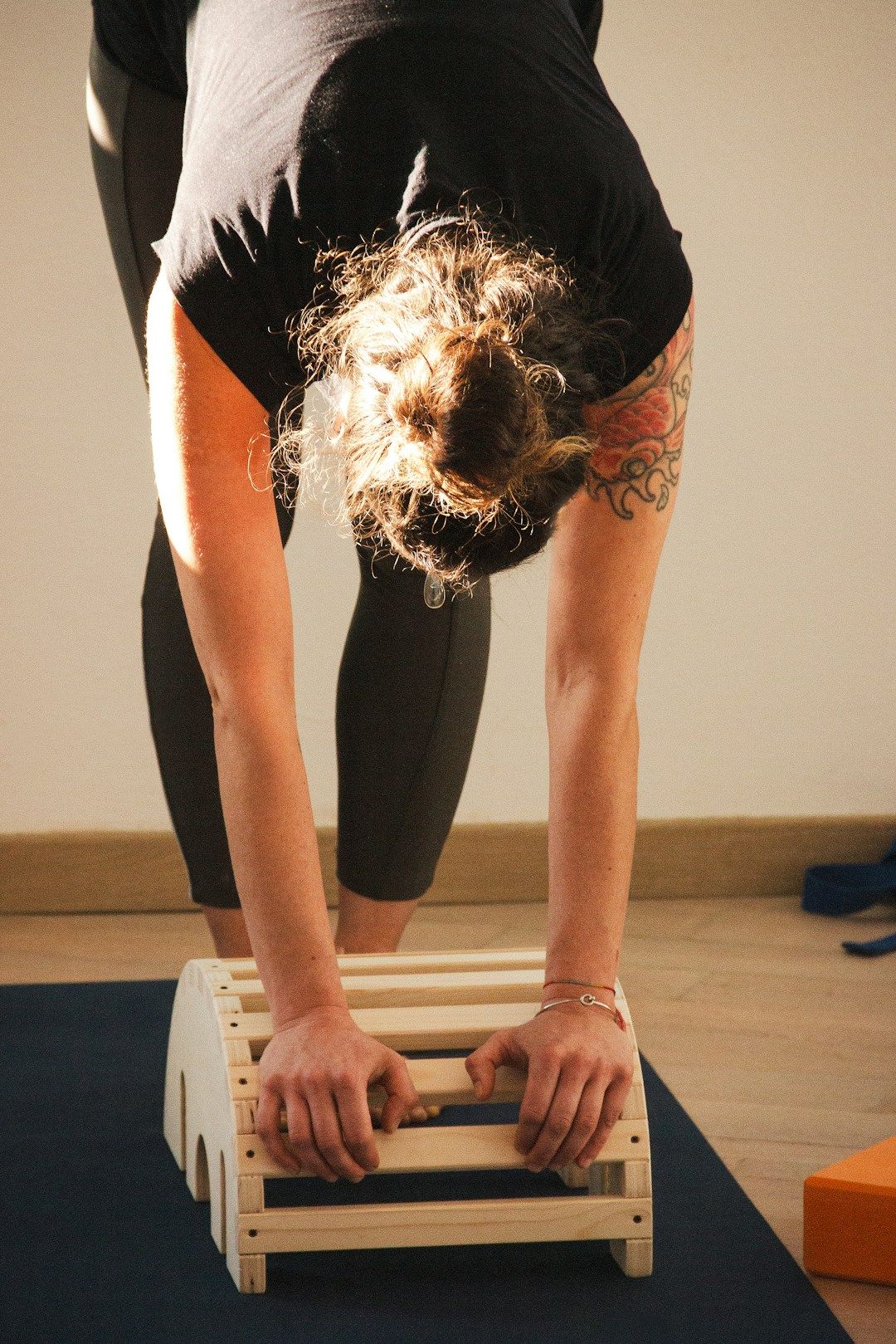 A yogi dressed in all black is seen in forward fold on their mat. They are using a wooden arch as yoga prop, elevating the ground to place their hands on.