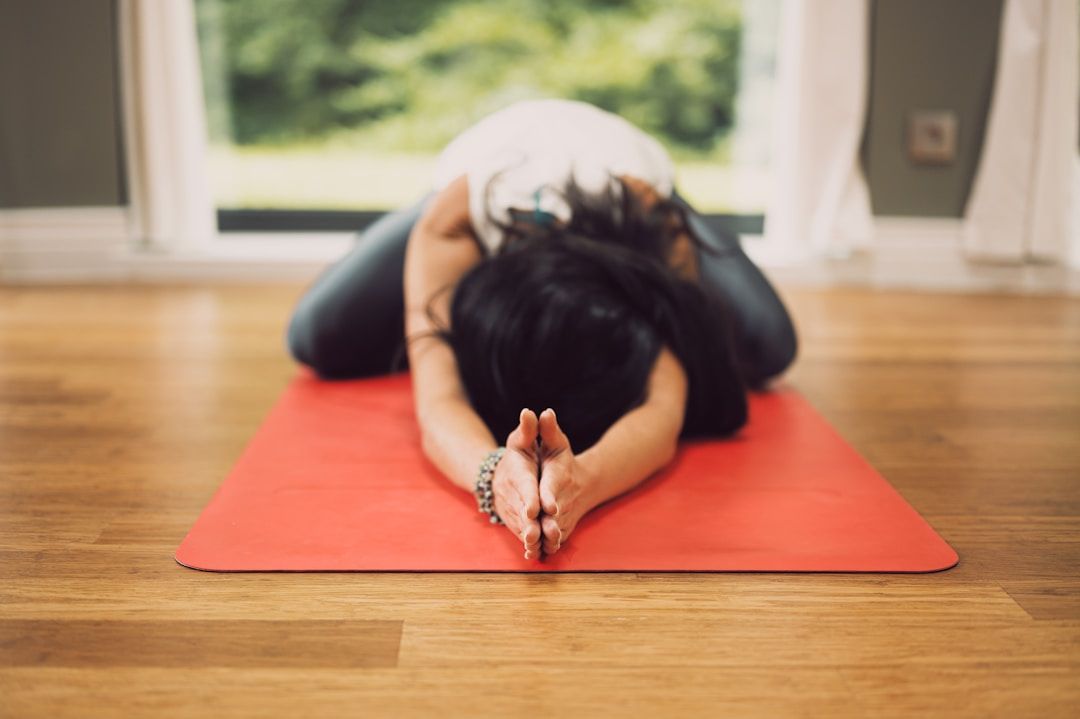a yogi is shown in Child's Pose with the hands touching in extension of the crown of the head. Their red mat is on a wooden floor with a blurred window behind them.