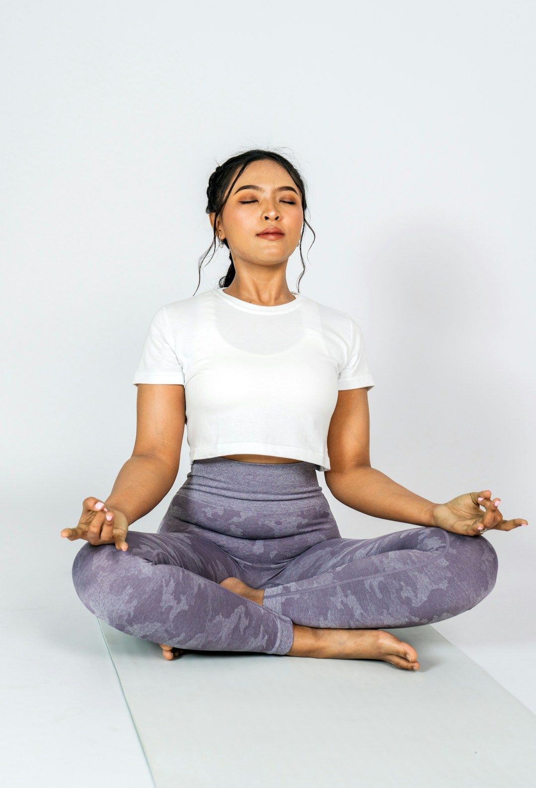 A yogi sits on a white mat in an all-white setting, wearing purple yoga pants and a white crop top. Their legs are crossed in front of them and their hands are resting on their knees, palms facing upward, thumbs and index fingers pressing gently together. Their eyes are closed and their dark hair is bound behind their head.