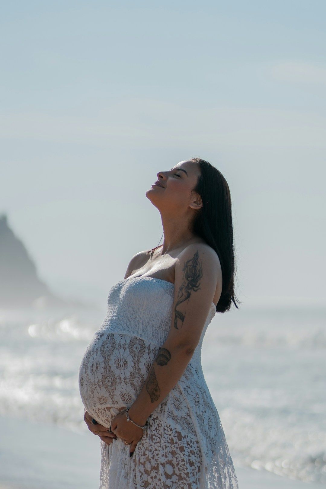 A pregnant women in a white see through dress is pictured against a blurred coastline. Her brown hair is falling open onto her back as she’s leaning her head backward, eyes closed.