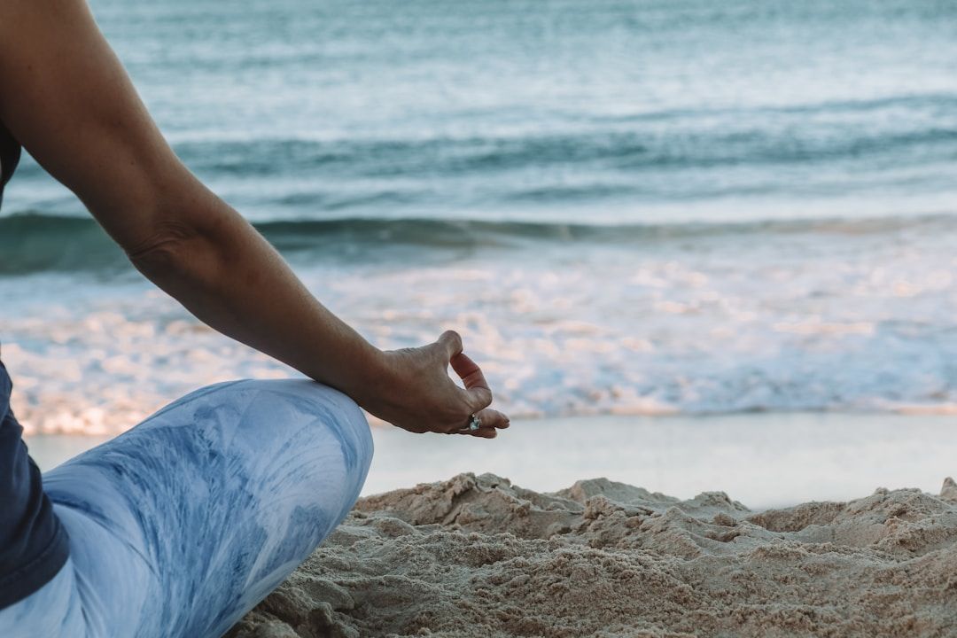 A yogis right knee and right arm are shown, sitting on the beach with legs crossed, thumb and index finger pressing gently together. In the background are gentle waves washing ashore.