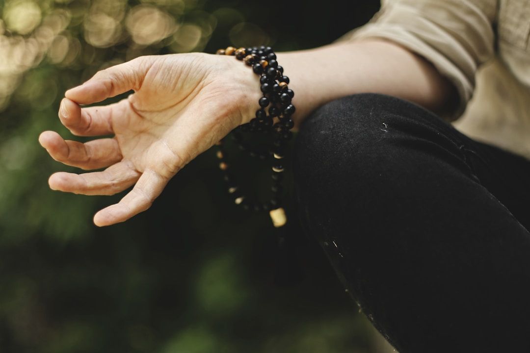 A knee dressed in a black leggings and a hand with lower arm are pictured, the arm dressed in a loosely rolled up khaki tunic with black and tiger’s eye male beads wrapped around the wrist. The lower arm is resting on the knee and thumb and index finger are gently pressing tighter. The background is dark, blurred trees