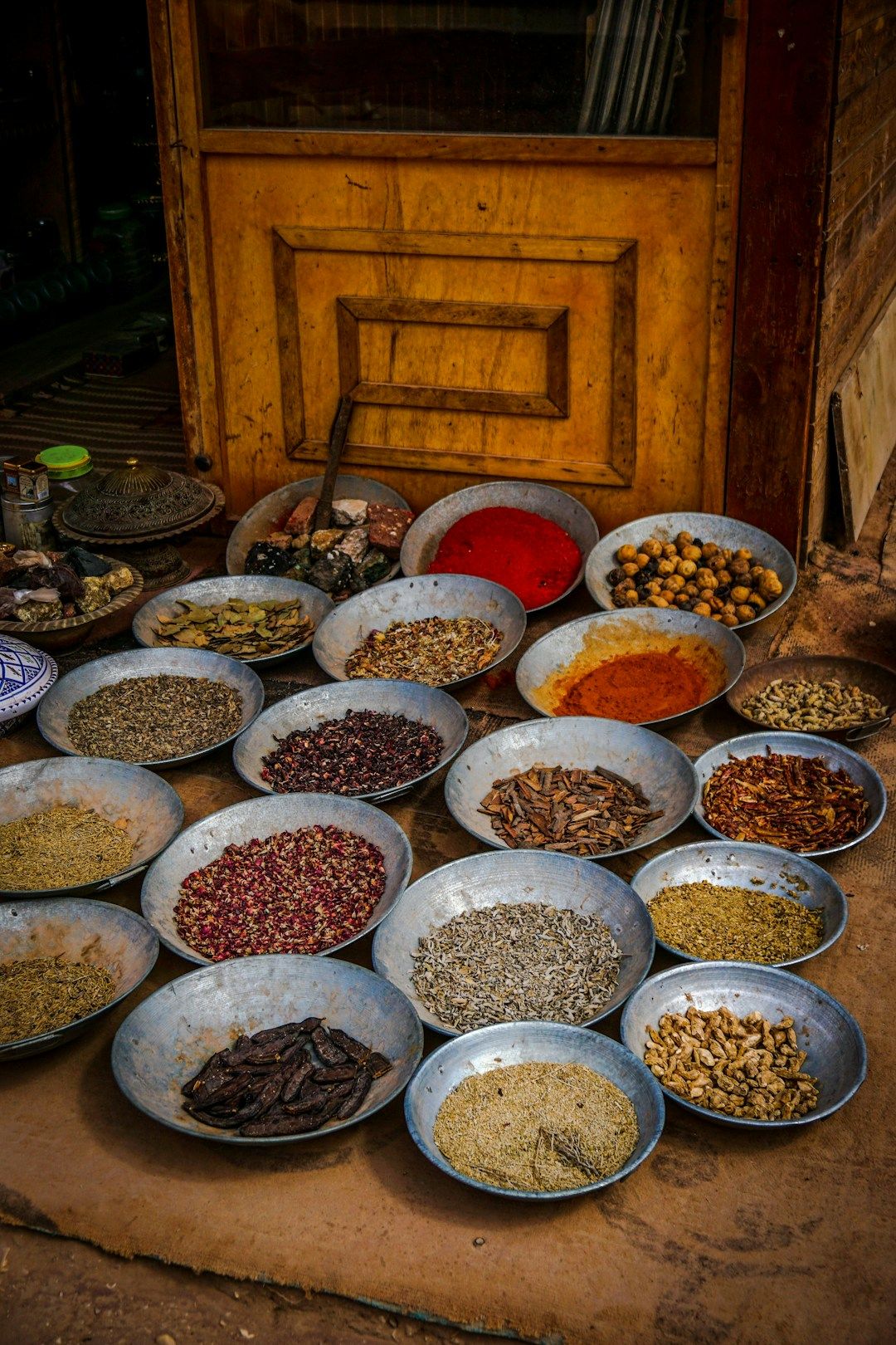 A wooden surface with multiple metal bowls holding different spices in their raw format. There’s also a wooden cupboard behind.