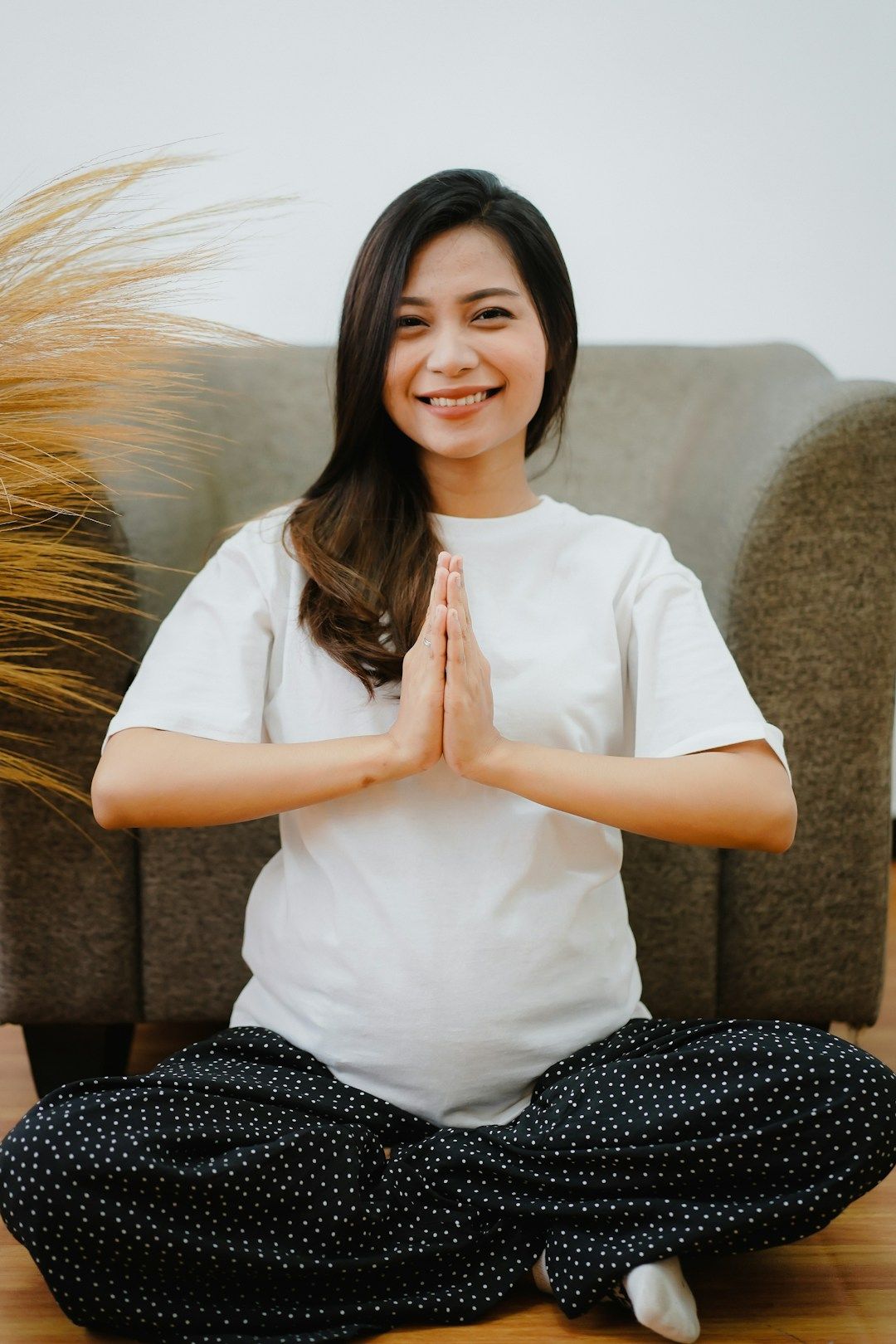 A pregnant yogi is sitting with their legs crossed in front of a green-grey sitting chair, with a beige plant reaching into the frame on the left hand side. They are wearing a white shirt and black pants with white dots, smiling at the camera. Their hands are gently pressing together in front of their chest and their brown hair falls open onto their back and right shoulder.