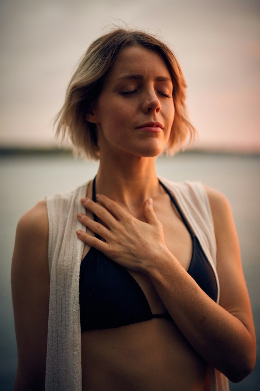 A woman bathed in the golden light of sunset stands with her eyes closed and her hand resting over her heart. She has short, wavy blonde hair and is wearing a deep purple top covered by a sleeveless, white, gauzy sweater. 