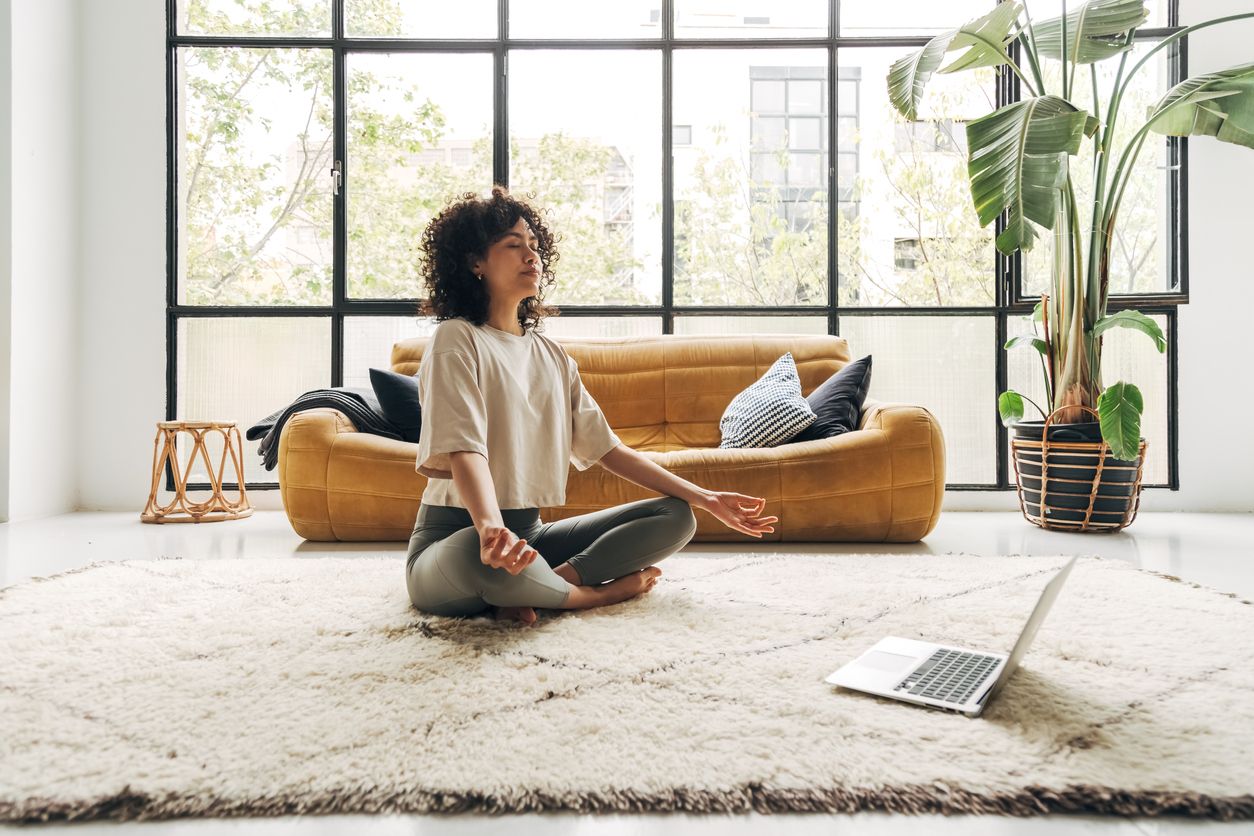 A woman sits on the ground in front of her laptop with her legs crossed. She's resting her hands on her knees facing upward, thumb and index fingers touching in a meditative pose. The woman is dressed in fitness clothes in earth tones, the background holds a large window front including an indoor palm tree and a yellow sofa.