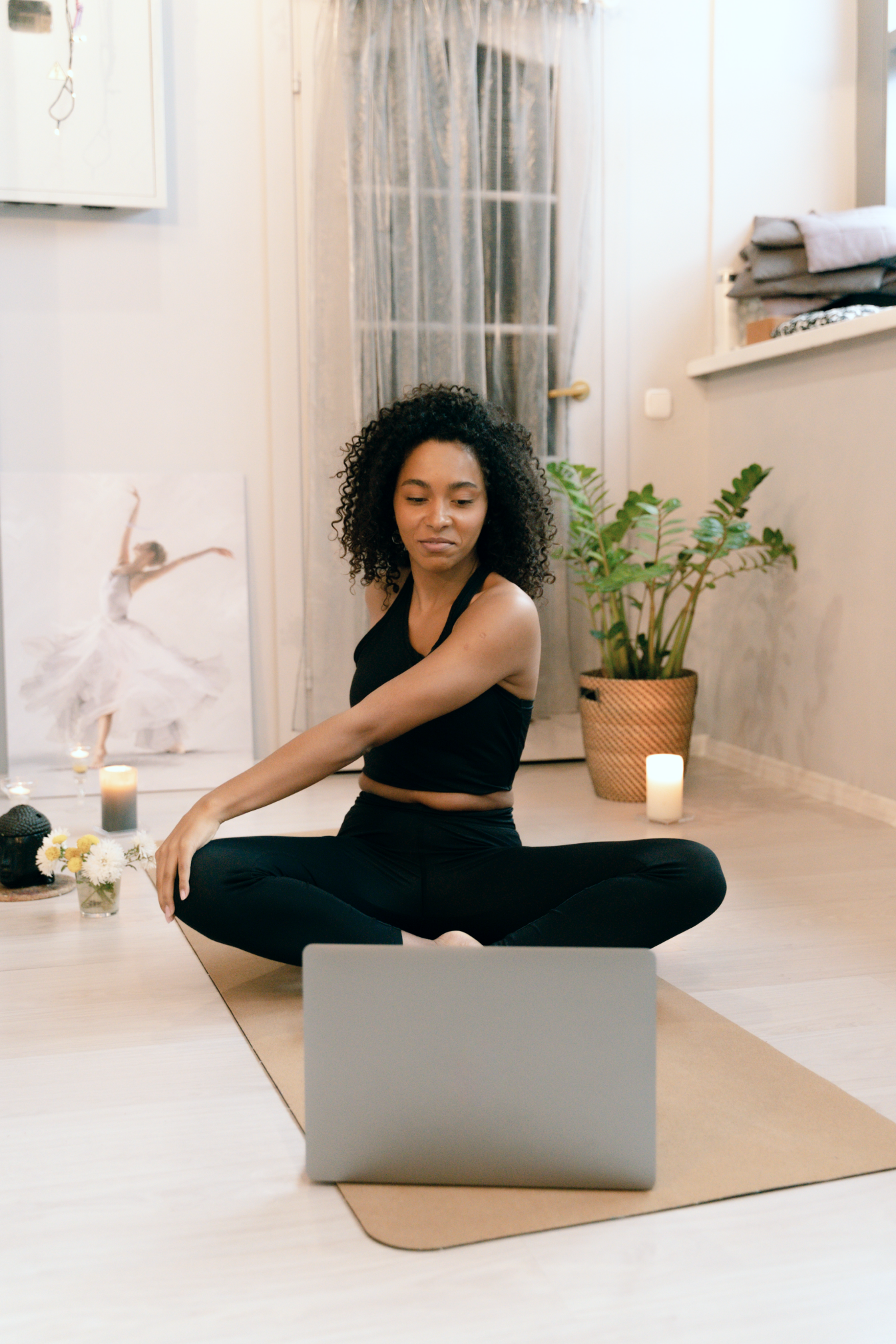 A woman sits on the ground in front of her laptop with her legs crossed. She has her left hand on the opposite knee in a stretch. She has curly hair and sits in front of a green plant. There are candles lit around her.