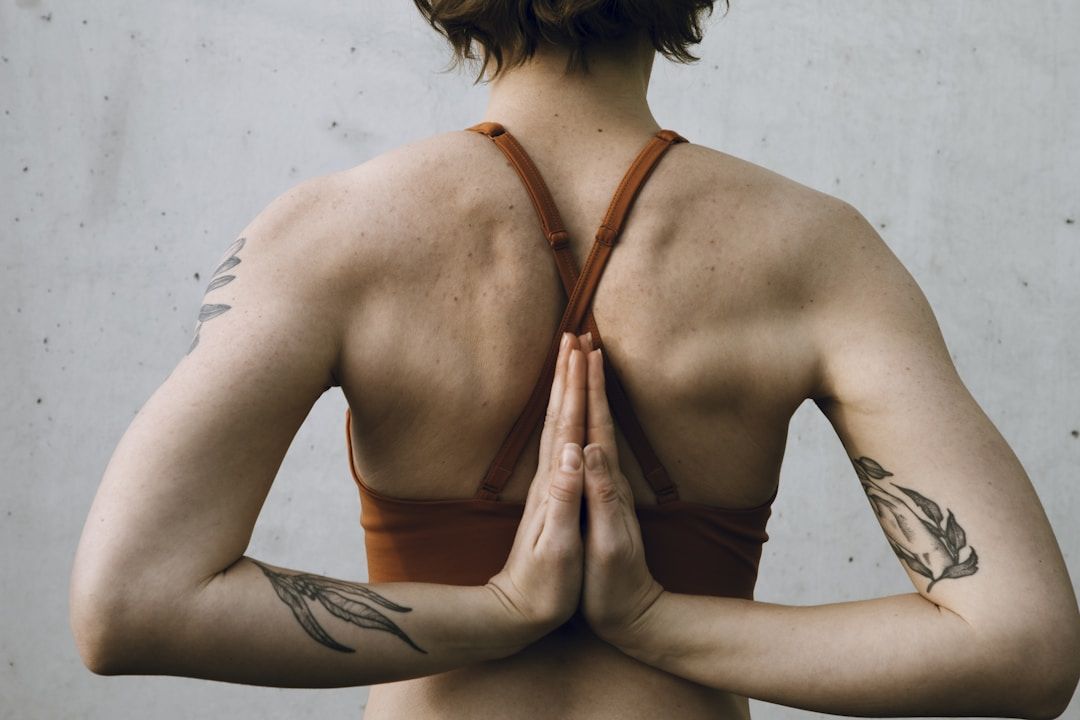 A yogi with short, brown hair is shown from behind, the palms of their hands meeting along their spine. They are wearing a mustard-coloured sports top and have tattoos of leaves on both arms. They are pictures against a light grey wall. 