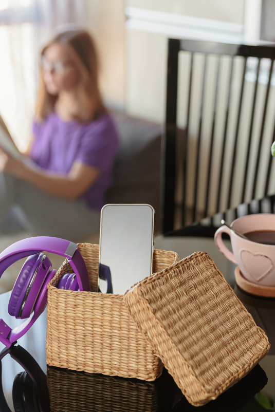 A wicker basket on a table holds purple over-ear headphones and a smartphone, with the basket lid beside it. In the blurred background, a person in a purple shirt sits on a sofa, and a pink mug of coffee rests on the table.