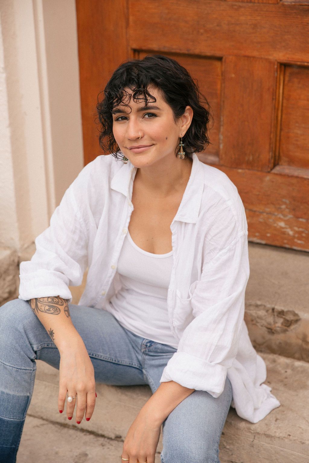 A woman sits on the ground in front of her laptop with her legs crossed. She has her left hand on the opposite knee in a stretch. She has curly hair and sits in front of a green plant. There are candles lit around her.