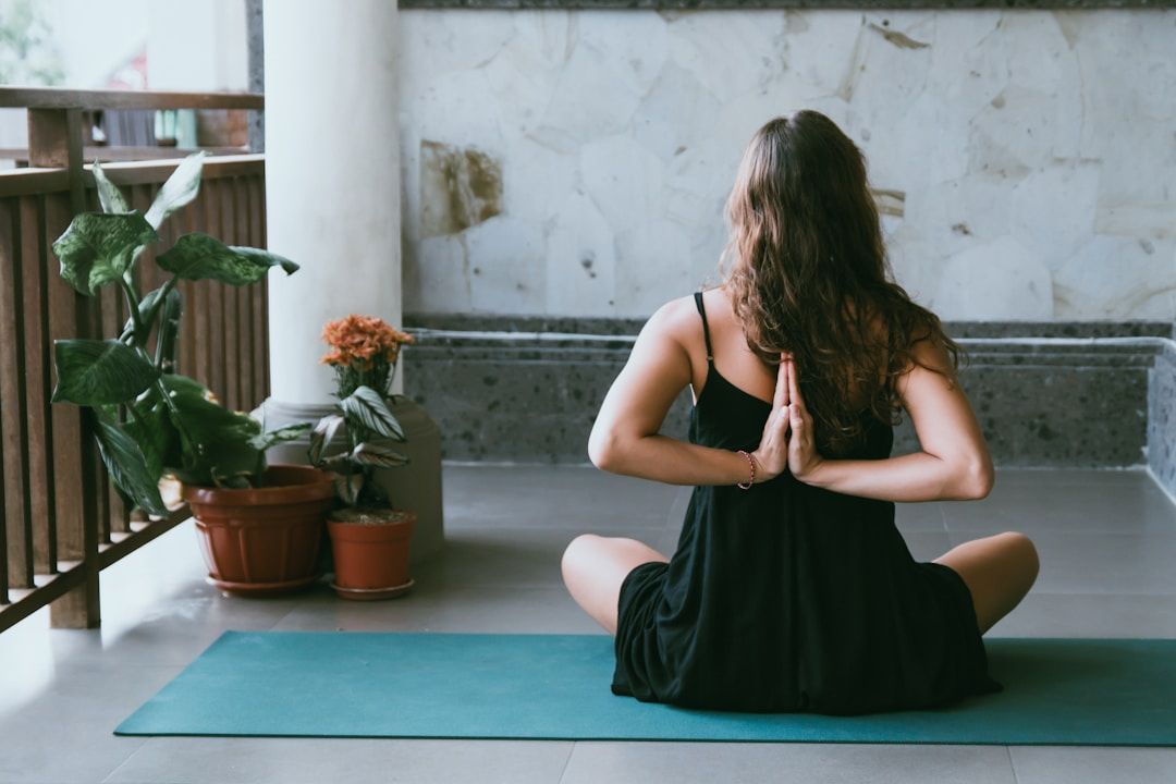 A woman sits on the ground in front of her laptop with her legs crossed. She's resting her hands on her knees facing upward, thumb and index fingers touching in a meditative pose. The woman is dressed in fitness clothes in earth tones, the background holds a large window front including an indoor palm tree and a yellow sofa.