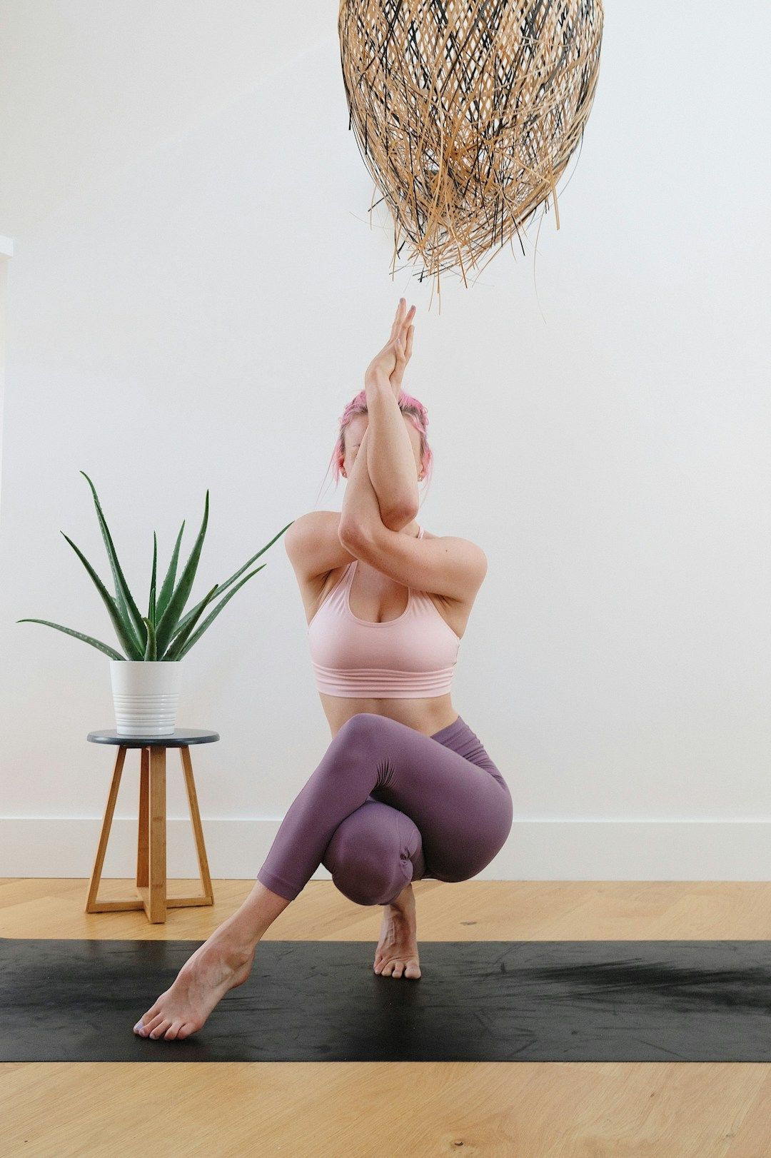 A woman is shown balancing on one leg, knee bent and sitbones touching the heel. The other leg is swung above the standing leg. the right arm threaded under the left, both arms are intertwined and raised in front of the face. She's wearing pink and purple-pastellgym clothes and practices on a black mat against a home background.