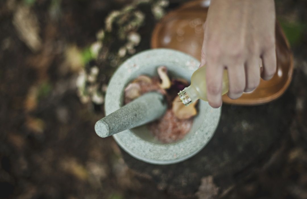 A hand is seen pouring a liquid into a mortar and pestle containing ingredients. The background is a soft, wooded area. 
