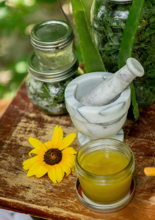 An image of a marble mortar and pestle with a prepared yellow mixture on a wooden carving board. There are jars of other preparations behind the mortar and pestle and there is a single sunflower on the board.