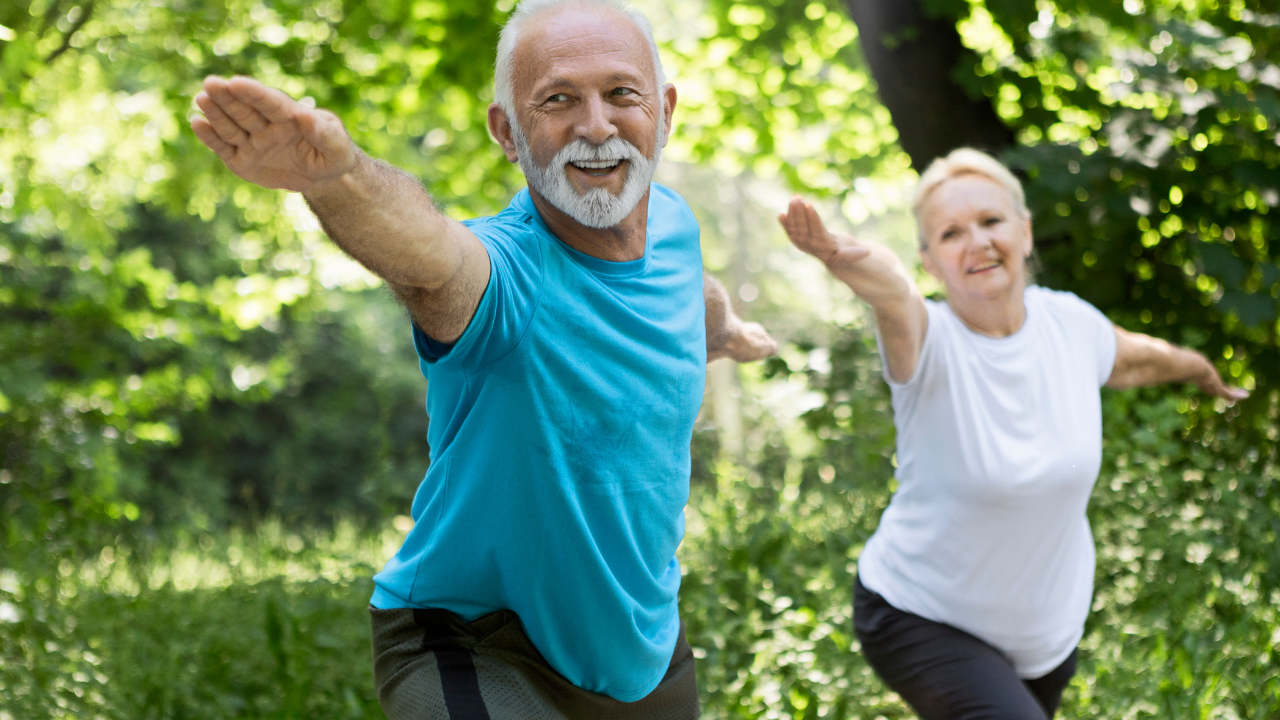 An older man and woman do yoga together in an outdoor setting. Sunlight filters softly through the green trees, as they both reach their arms forward. The man has short gray hair and a short gray beard and mustache. He is wearing a teal-colored short-sleeve shirt and dark gray pants. The woman also has gray hair, pulled back into a ponytail. She is wearing a white, short-sleeve athletic shirt and black leggings. Both are smiling. 