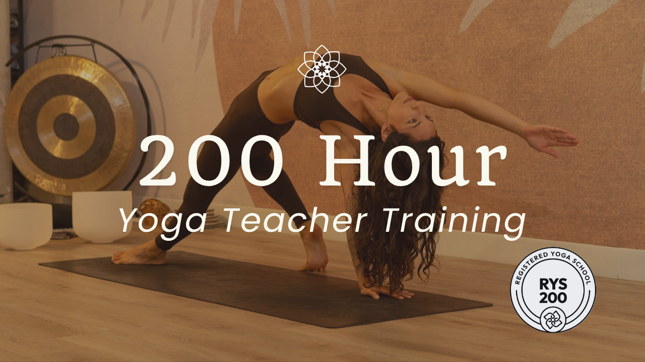 A yoga instructor in a black outfit assists a student in adjusting her posture during a yoga class. The student, also dressed in black, is in a twisting pose with hands in prayer position. Other participants practice poses in the background in a softly lit studio.