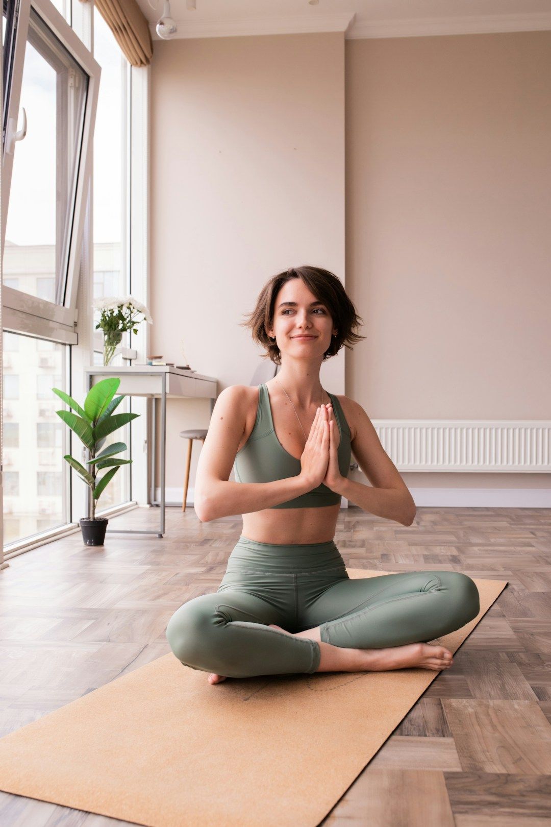 A woman with brown, chin-long hair is pictures on a beige yoga mat in a room with wooden floors and light-brown walls. There’s a desk and plant in the background, as well as a floor length window front on the left. The woman is wearing a khaki yoga set and looking to the left of the camera. The palms of her hands are gently pressing together in front of her chest and her legs are crossed in front of her. 