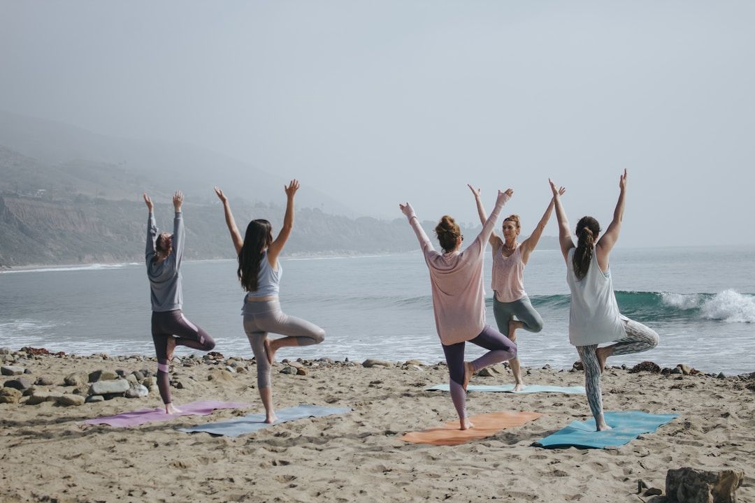 A group of woman is shown on the Bach, all in Tree Pose with their right leg raised and gently pressed against the left thigh. Their arms are raised overhead, reaching upward. 