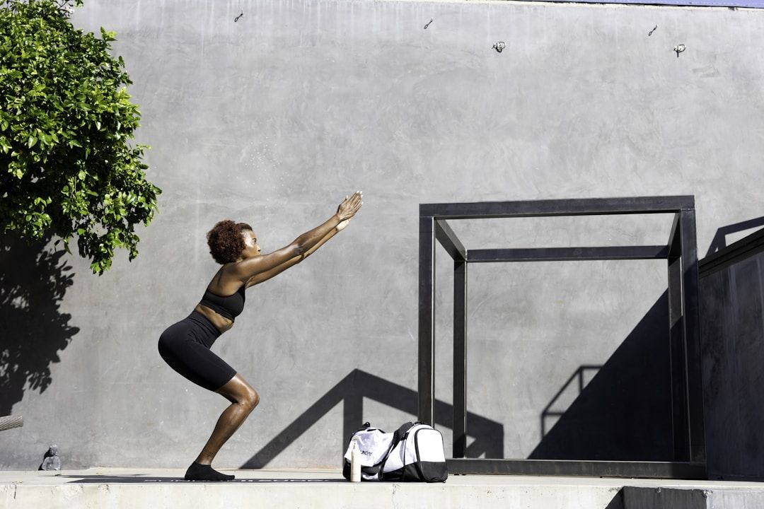 A woman sits on the ground in front of her laptop with her legs crossed. She's resting her hands on her knees facing upward, thumb and index fingers touching in a meditative pose. The woman is dressed in fitness clothes in earth tones, the background holds a large window front including an indoor palm tree and a yellow sofa.